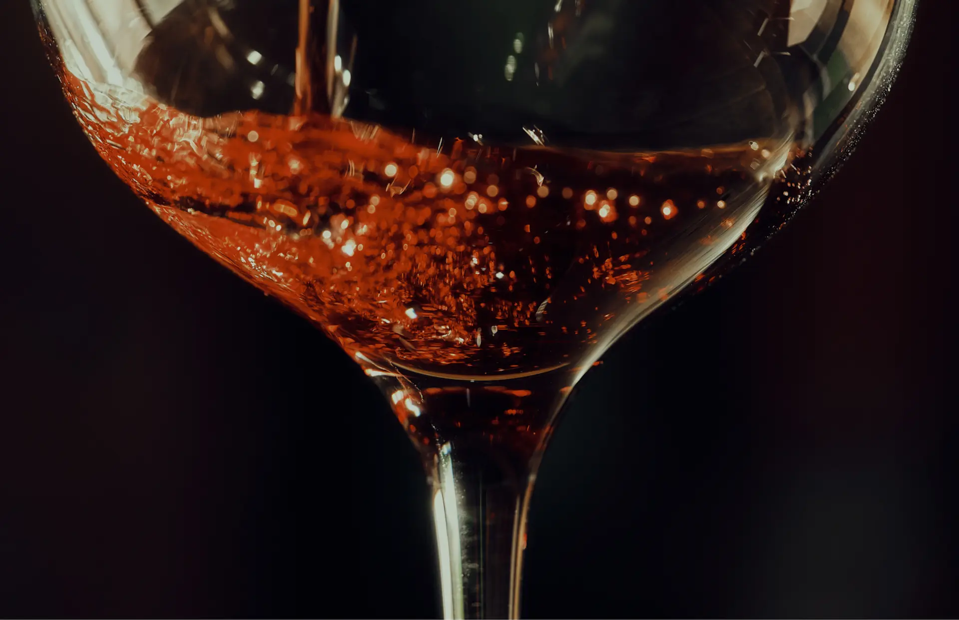 Close-up of red wine being poured into a glass, with amber tones and sparkling reflections against a dark background.