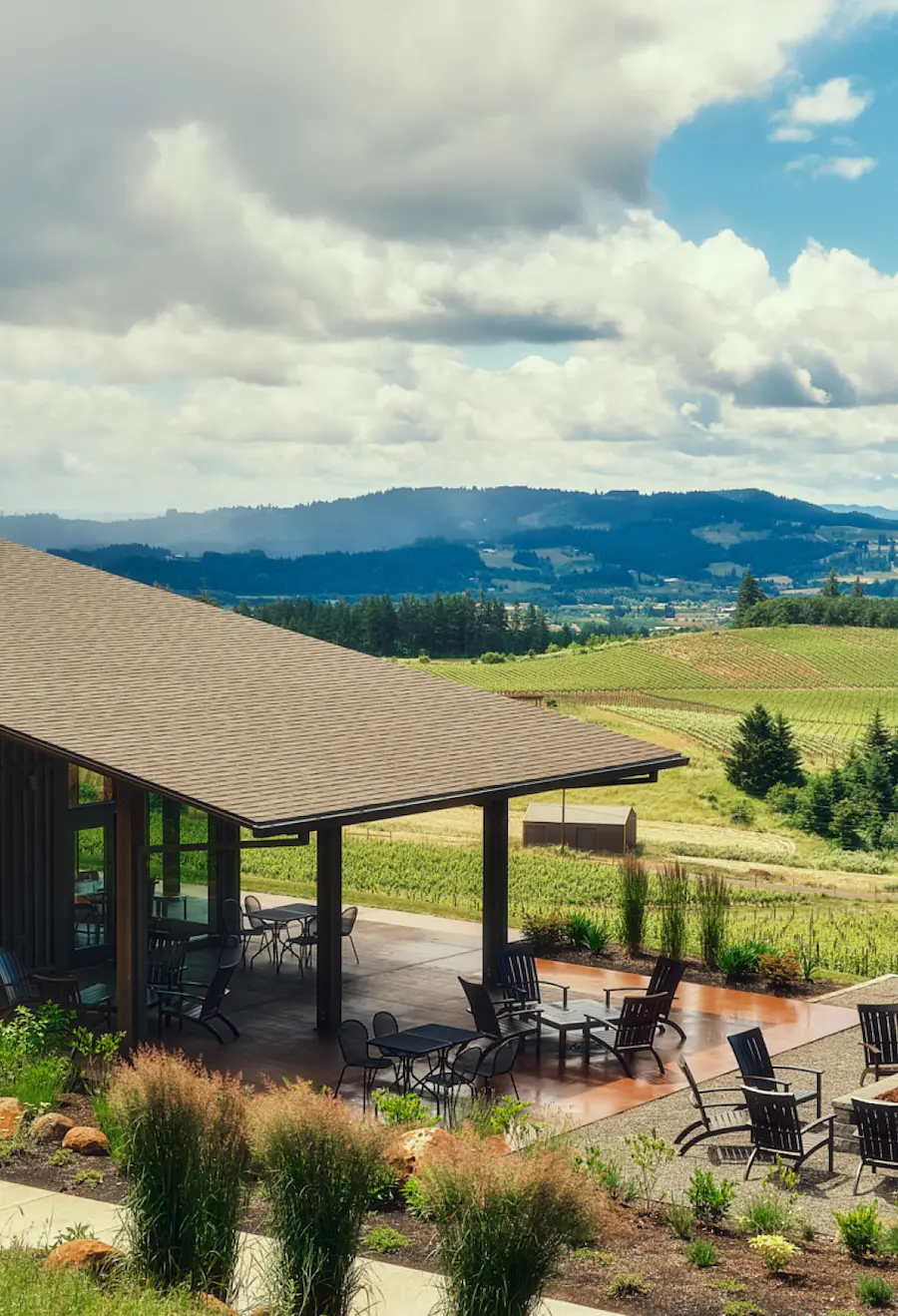 A modern winery with an outdoor patio and fire pit overlooking rolling vineyards and hills under a partly cloudy sky in Oregon.