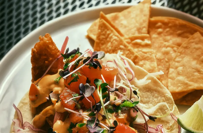 A crispy fish taco from Dos Mundos with fresh toppings, served with tortilla chips, lime, and radish on a white plate.