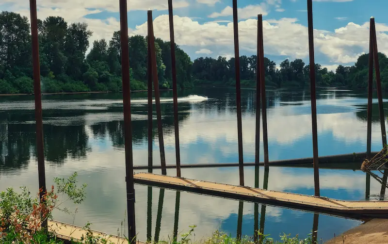A calm river reflecting the sky, with wooden docks and tall poles, surrounded by lush green trees.