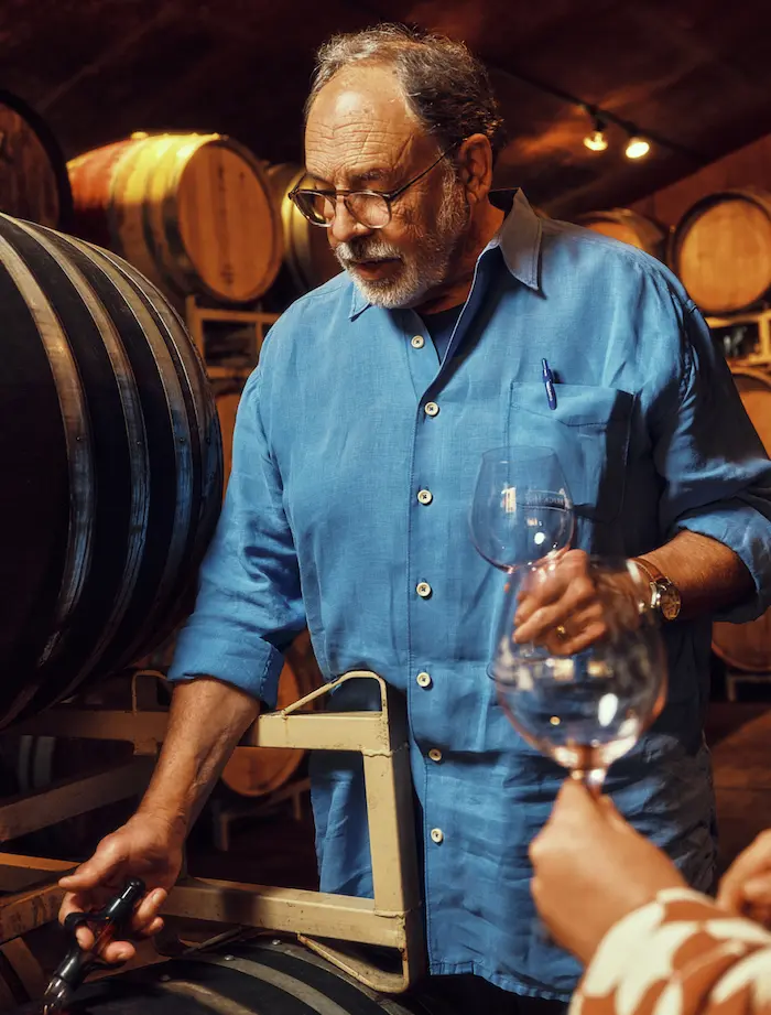 A winemaker in a blue shirt pours wine from a barrel in a dimly lit cellar lined with wooden casks.
