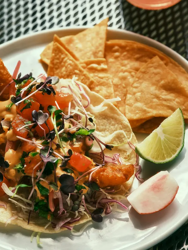 A crispy fish taco from Dos Mundos with fresh toppings, served with tortilla chips, lime, and radish on a white plate.