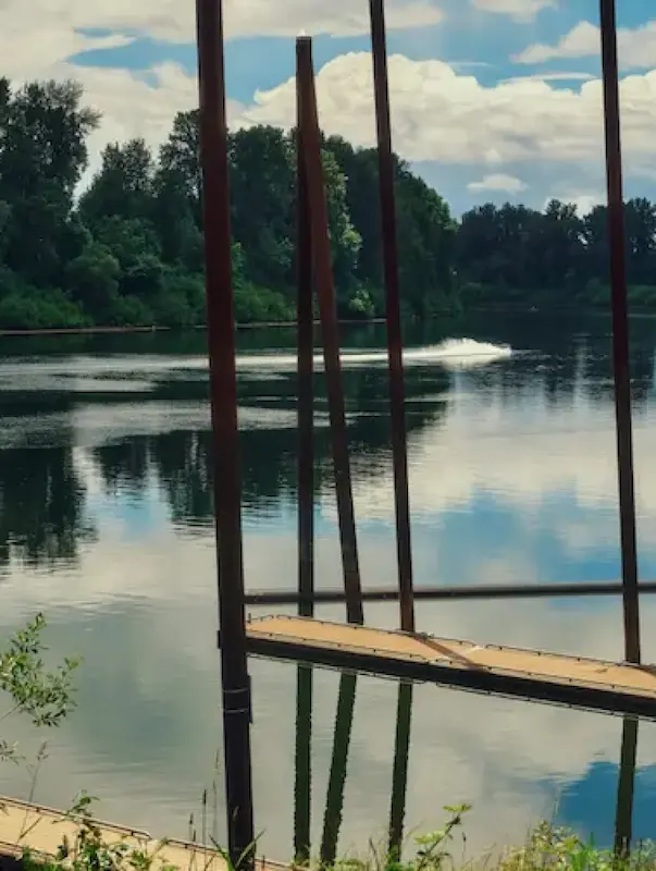 A calm river reflecting the sky, with wooden docks and tall poles, surrounded by lush green trees.