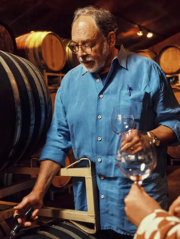A winemaker in a blue shirt pours wine from a barrel in a dimly lit cellar lined with wooden casks.