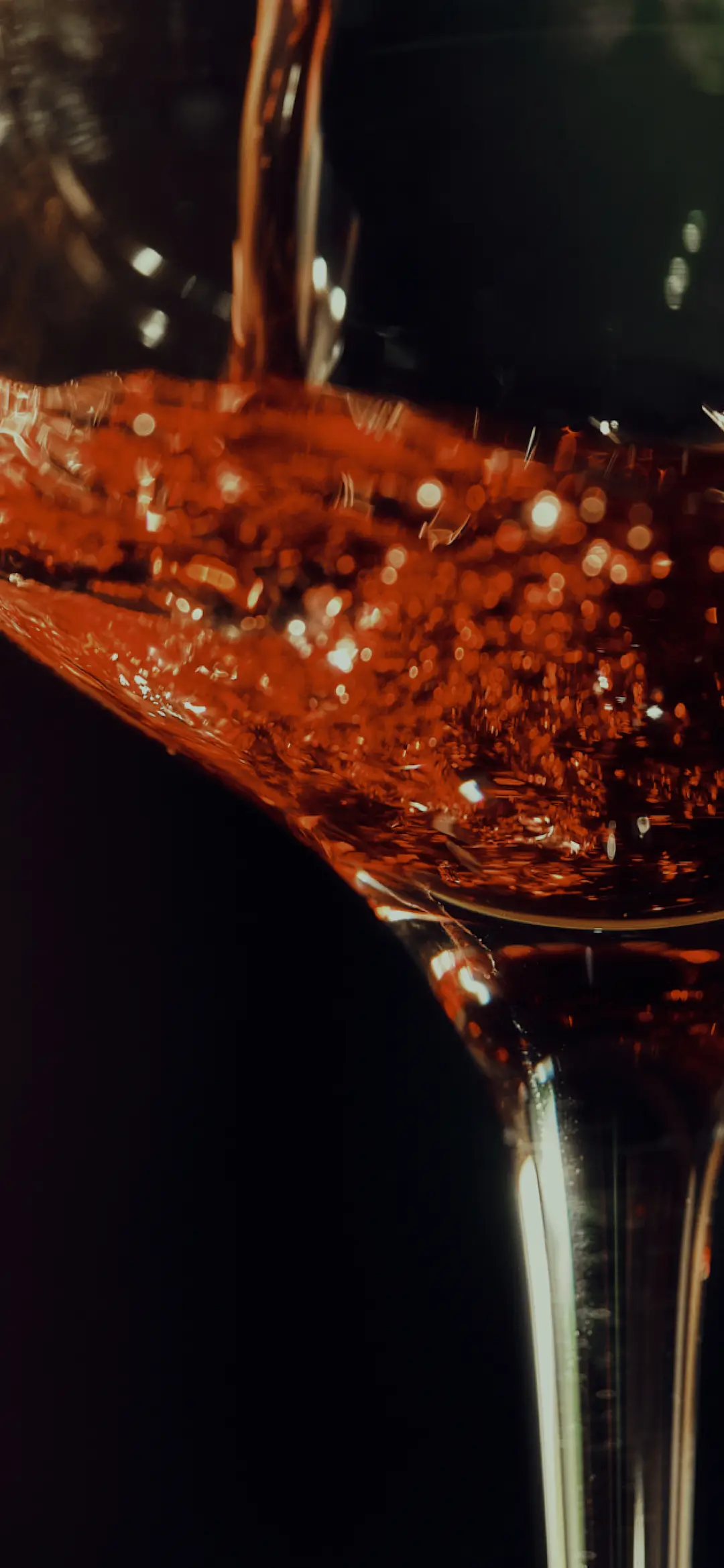 Close-up of red wine being poured into a glass, with amber tones and sparkling reflections against a dark background.